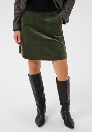 Femme portant une jupe en velours côtelé vert olive, des bottes noires montantes au genou et un blazer foncé, une main dans la poche.