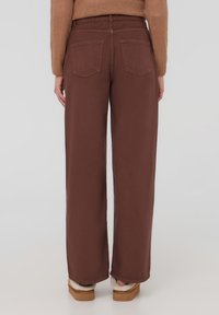 Pantaloni larghi marroni con vita alta, due tasche posteriori e una superficie liscia. Indossati con scarpe slip-on pelose di colore chiaro.