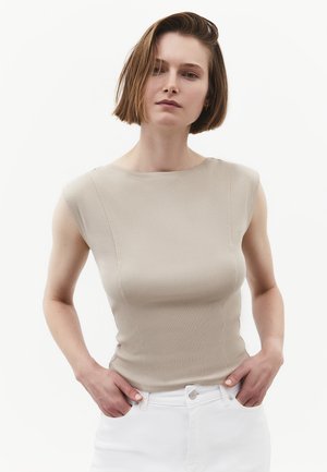 Frau mit kurzen braunen Haaren, die ein ärmelloses beiges Oberteil und weiße Hose trägt, steht mit den Händen in den Taschen vor einem einfarbigen Hintergrund.