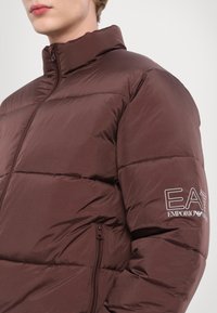 Brūna pūkā jaka ar augstu apkakli, quilted dizainu un rāvējslēdzēja kabatu. Iespējama redzama zīmola izcelšana uz piedurknes baltā krāsā.