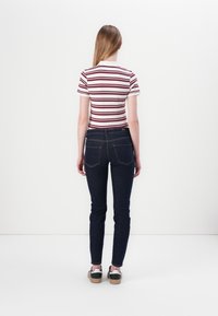 Γυναίκα με μακριά μαλλιά που φοράει λευκό και μπορντό ριγέ κοντομάνικο μπλουζάκι, σκούρα στενά τζιν και μαύρα-άσπρα αθλητικά παπούτσια, γυρισμένη πλάτη.