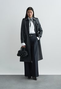 Trench-coat en cuir noir avec poignets blancs contrastants, associé à une chemise blanche et une cravate grise. Pantalon noir taille haute et petit sac noir avec accents argentés.