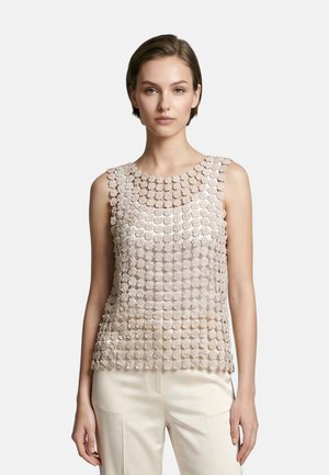 Donna che indossa un top senza maniche in pizzo floreale beige sopra un camisole abbinato e pantaloni color crema, in piedi davanti a uno sfondo semplice.