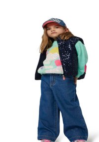 Denim wijde-broek in combinatie met een mintgroen gebreid sweater met patroon en een marineblauwe pailletten vest. Een blauwe pet met een roze rand maakt de outfit compleet.
