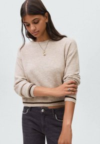 Maglione a maglia beige chiaro con scollo rotondo, polsini a costine e strisce marrone scuro all'orlo, abbinato a jeans scuri con dettagli a borchie.