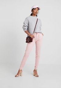 Lichtgrijze gebreide trui, roze joggers, een roze pet en een zwarte crossbodytas met rode accenten; draagt gouden hakken.