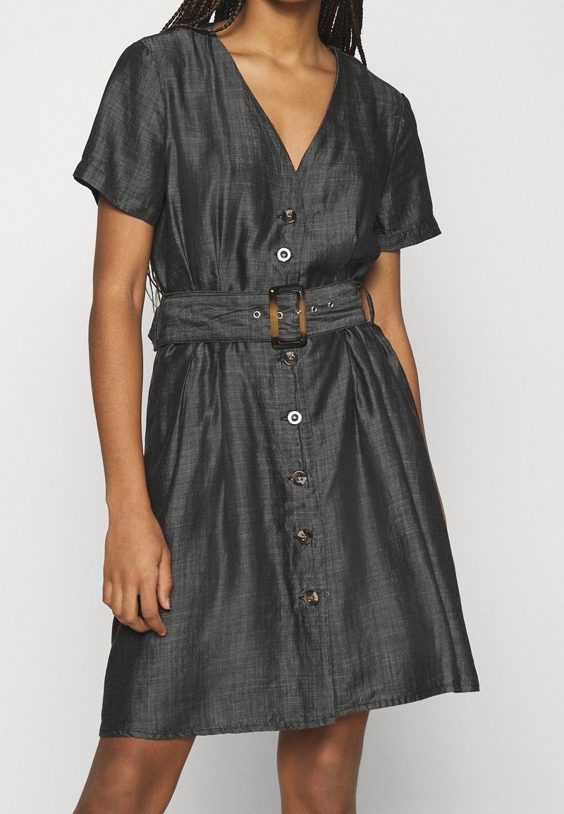 Femme portant une robe noire à manches courtes avec boutons sur le devant, un décolleté en V et une large ceinture avec une boucle rectangulaire à la taille.