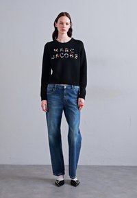 Pulover negru cu "MARC JACOBS" în litere multicolore, asociat cu blugi drepti de culoare albastră deschisă și pantofi negri. Fundal simplu.