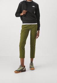 Mustapaita, jossa on logo, oliivinvihreät nyöripohjat, ja harmaavihreät vaelluskengät, joissa on oranssit yksityiskohdat. Reppu kantaa olkapäällä.