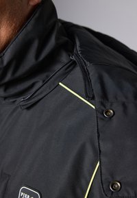 Chaqueta negra impermeable con un alto cuello, ribetes amarillos, un bolsillo con cremallera y detalles de botones a presión en el hombro.