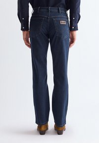 Vue arrière d'une personne portant un jean Wrangler bleu foncé, une chemise à manches longues foncée et des chaussures à talons marron, debout sur fond blanc.