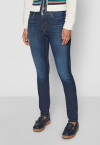 Jeans skinny azul oscuro de denim, con un diseño clásico de cinco bolsillos y costuras en contraste; usados con mocasines marinos.