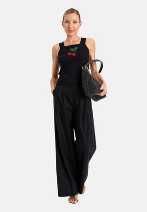 Donna che indossa un top nero senza maniche con motivo di ciliegie rosse, pantaloni neri a gamba larga, tiene una grande borsa intrecciata nera, in piedi su sfondo bianco.
