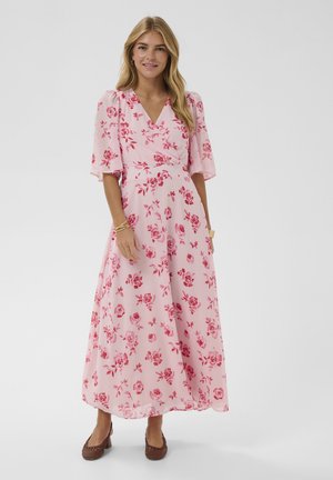 Femme aux longs cheveux blonds portant une robe portefeuille fleurie rose et des ballerines brunes, debout devant un fond blanc.