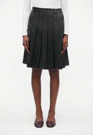 Kobieta w ciemnoszarej plisowanej spódnicy do kolan, białej bluzce z długim rękawem oraz ciemnoczerwonych płaskich butach stojąca na białym tle.