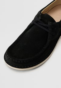 Juodos suede batai su apvaliu priekiu, kontrastingomis siūlėmis ir juodais raišteliais. Padas yra šviesios spalvos ir šiek tiek pakeltas.