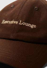 Rjav bombažni pokrivalo z vezenim napisom "Executive Lounge" v svetlo bež barvi, z gladko teksturo in ukrivljenim visokom.