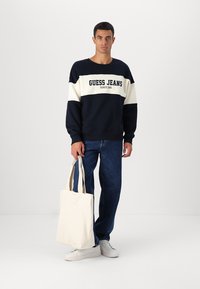 Marineblauwe en crème sweatshirt met de tekst "GUESS JEANS", gecombineerd met blauwe spijkerbroek en witte sneakers. Houdt een crème tote bag vast.