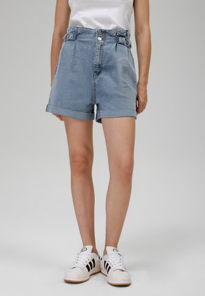 Shorts de mezclilla azul claro con una cintura alta, puños enrollados y cierre frontal de botón. Combinados con zapatillas blancas que tienen rayas negras.