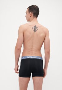Crne bokserice s plavim Nike pojasom, imaju prilagođeni dizajn i minimalno šivanje. Pogled s leđa prikazuje tetovažu na gornjem dijelu leđa.