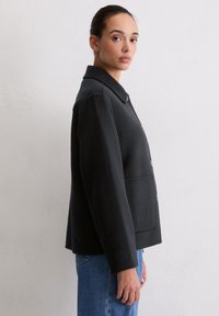 Czarna kurtka z mieszanki wełny z kołnierzem, zapięciem na guziki i boczną kieszenią, o prostym kroju i gładkiej fakturze.