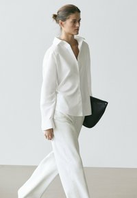 Femme marchant à l'intérieur, portant une chemise blanche boutonnée et un pantalon blanc, tenant une pochette noire avec les cheveux attachés.