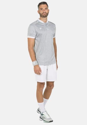 Uomo che indossa una polo grigia a fantasia, pantaloncini bianchi, un braccialetto e sneakers bianche, in piedi davanti a uno sfondo semplice.