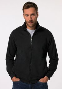 Chaqueta negra con cremallera fabricada en tejido suave, con un cuello alto, que cuenta con bolsillos laterales y puños acanalados. Diseño simple y estilizado.