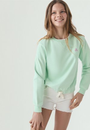 Adolescente sonriente que lleva una sudadera verde claro con un pequeño logo de calavera rosa y pantalones cortos blancos, de pie sobre un fondo liso.