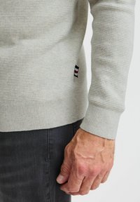 Pull en tricot gris clair avec des poignets et un ourlet côtelés, arborant une petite étiquette logo tricolore sur le côté. La texture est subtile et tissée.