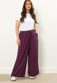 Śliwkowe spodnie o szerokich nogawkach z teksturowanym wykończeniem, elastycznym pasem i detalem sznurka. W połączeniu z prostą białą koszulką z krótkim rękawem.