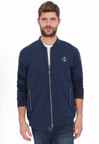 Marineblå bomberjakke med lynlås foran, to lynlåslommer i siderne, ribbet manchetter og et lille ankerlogo på brystet. Letvægtsstof.