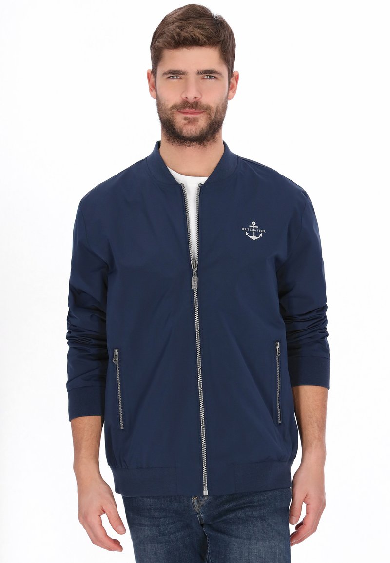 Marineblå bomberjakke med lynlås foran, to lynlåslommer i siderne, ribbet manchetter og et lille ankerlogo på brystet. Letvægtsstof.