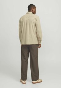 Camicia a maniche lunghe beige con colletto abbottonato, tessuto testurizzato e pieghe posteriori, abbinata a pantaloni marroni e sneakers neutre.