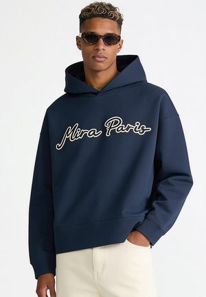 Junger Mann trägt dunkelblauen Kapuzenpullover mit dem Text "Mira Paris" und Schildpatt-Sonnenbrille, steht mit einer Hand in der Tasche.