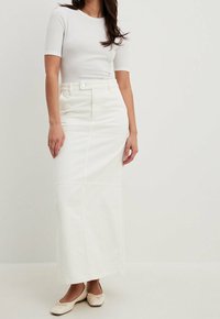 Jupe maxi en denim blanc avec une taille ajustée, fermeture à boutons sur le devant et détails de panneaux cousus, associée à un haut blanc ajusté et des chaussures légères.