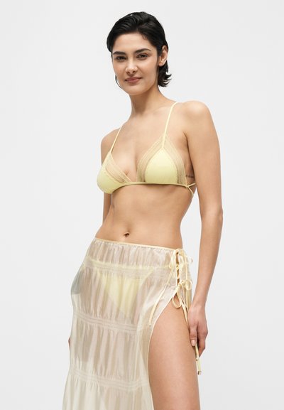 Γυναίκα με κοντά σκούρα μαλλιά, που φοράει ένα κίτρινο σουτιέν με δαντέλα και μια διάφανη κρεμ φούστα με κορδόνι στο πλάι, σε λευκό φόντο.