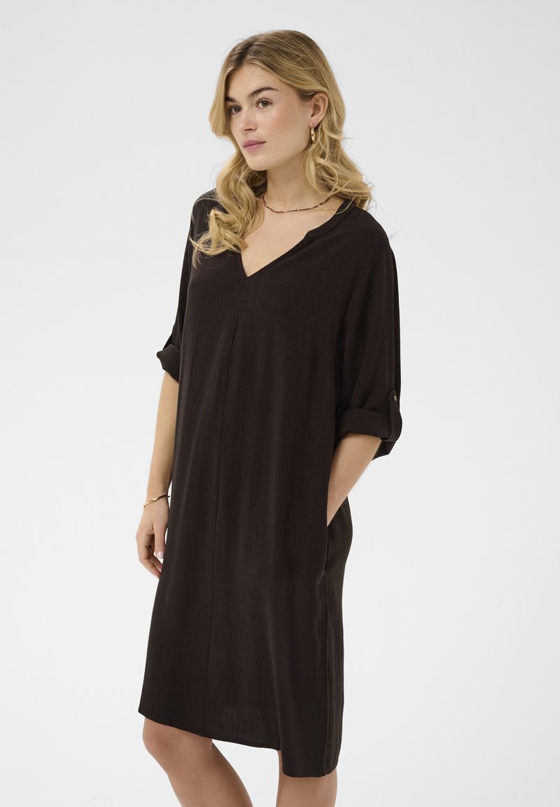 Femme blonde portant une robe noire ample mi-genoux avec les manches retroussées, debout avec une main dans la poche contre un fond uni.