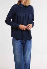 Blusa a maniche lunghe blu navy con colletto e dettagli arricciati sul petto. Tessuto liscio, abbinata a jeans di denim blu chiaro.