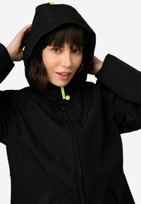 Veste à capuche noire avec une fermeture éclair intégrale, accentuée d'un vert vif sur la tirette de la fermeture et la doublure intérieure ; tissu lisse et léger.