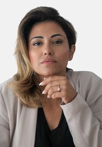 Donna con capelli castani ondulati, che indossa un blazer beige chiaro sopra una maglietta nera, mentre tiene in mano un anello d'argento con una perla.