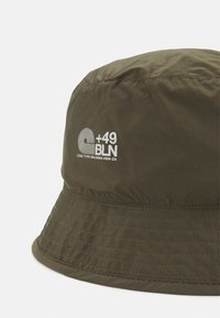 Chapéu de balde verde oliva feito de tecido leve. Possui uma copa arredondada, aba larga e plana, e um logótipo branco na parte da frente.