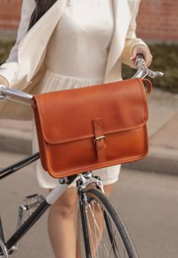 Brun læder messenger taske med en frontlåg og spænde lukning, hængende på en cykels styr; glat tekstur og rene linjer.