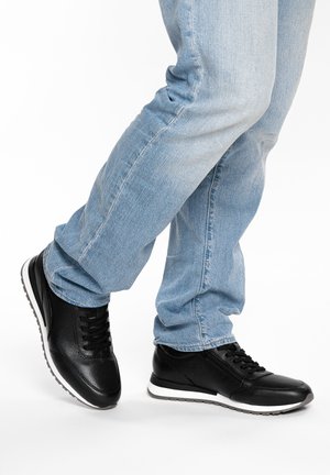 Person trägt hellblaue Jeans und schwarze Sportschuhe, steht mit einem leicht angehobenen Fuß auf weißem Hintergrund.