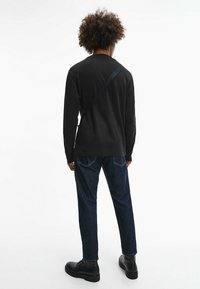 Svart långärmad topp, denimjeans, svarta snörkängor och en justerbar rem över axeln, sedd bakifrån mot en vit bakgrund.