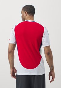 Tricou sportiv roșu și alb cu textură din plasă, mâneci scurte și accente albastre; are un panou roșu rotunjit pe spate.