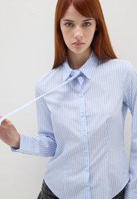 Camicia a righe blu e bianche con colletto a punta, dalla trama liscia e maniche lunghe, dotata di una fila di bottoni sul davanti.