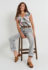 Top de manga corta con escote en V en gris claro con un patrón de hojas coloridas; vaqueros de mezclilla claros de cintura alta; mules de madera marrones con tacón.