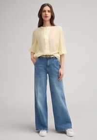 Blusa gialla leggera con maniche corte e larghe e una chiusura a bottoni, abbinata a jeans in denim blu larghi e sneakers bianche.