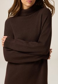 Suéter marrón de punto con cuello alto y puños acanalados. Textura suave, corte relajado. Brazos cruzados, cubriendo parcialmente el cuerpo.